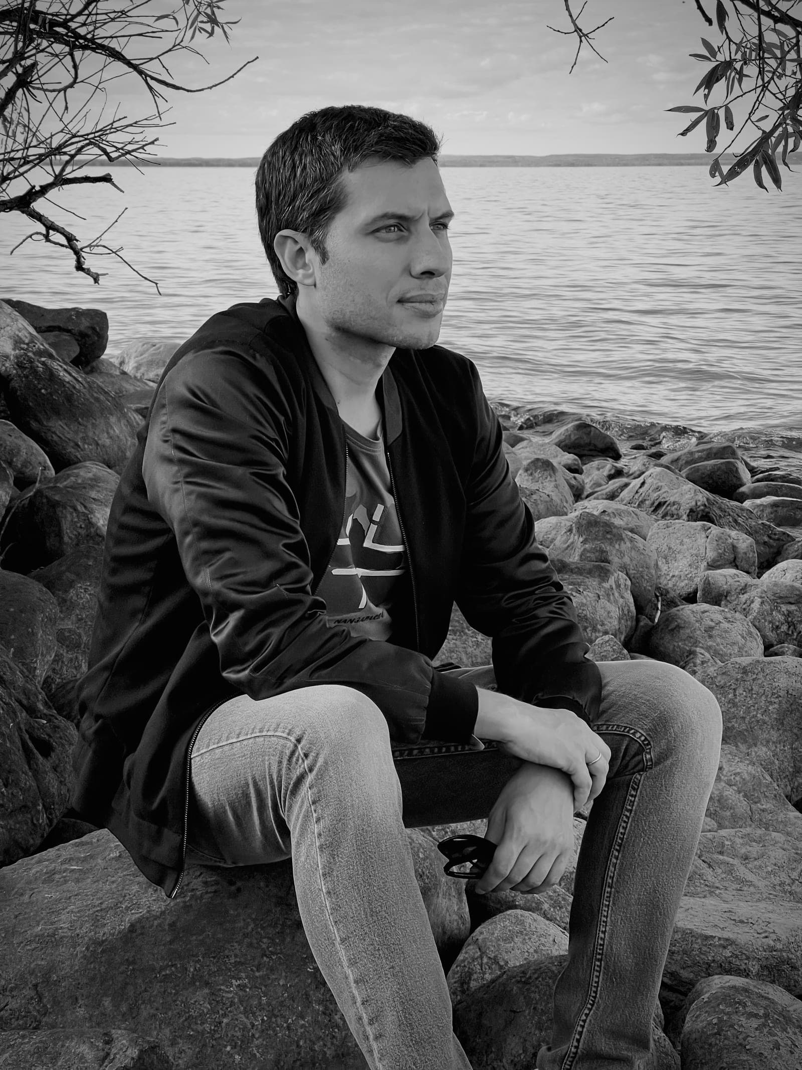 Black-and-white portrait of Nazar Khomyshyn sitting on rocks beside a lake, in a dark jacket and jeans, looking out across the water.